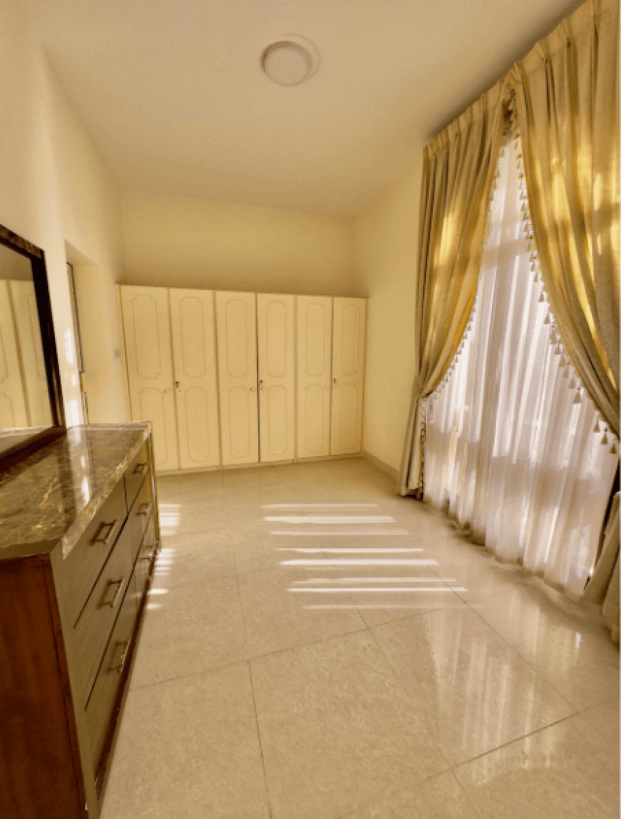 Bright room with a wooden dresser, cream-colored wardrobes, and a window with long, light-colored curtains. Sunlight casts shadows on the tiled floor.